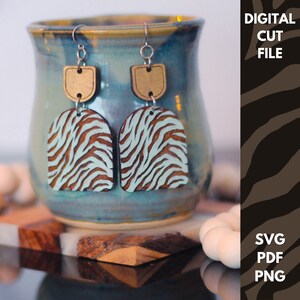 Könnte beinhalten: Ein Paar Holzohrringe mit einem hellblauen und braunen Zebra-Muster. Die Ohrringe haben ein goldfarbenes Oberteil und silberne Haken.