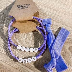 May include: Two lavender-colored bracelets with white letter beads spelling "Skyla" and "Roxy". A matching lavender hair tie is also included. The bracelets and hair tie are displayed on a wooden surface, with a brown tag that reads "Boundless Autumn".