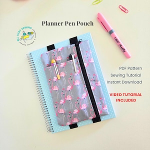 May include: A gray fabric pen pouch with a pink flamingo print and a black zipper, attached to a light blue spiral-bound notebook with white polka dots. The pouch features two vertical pockets holding pens, with text reading Planner Pen Pouch.