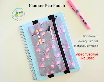 May include: A gray fabric pen pouch with a pink flamingo print and a black zipper, attached to a light blue spiral-bound notebook with white polka dots. The pouch features two vertical pockets holding pens, with text reading Planner Pen Pouch.
