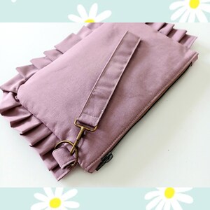 May include: A mauve fabric clutch bag featuring a ruffled edge detail along the side. The rectangular pouch includes a matching detachable wrist strap secured with a bronze-toned metal lobster clasp and a black zipper closure along the top edge.