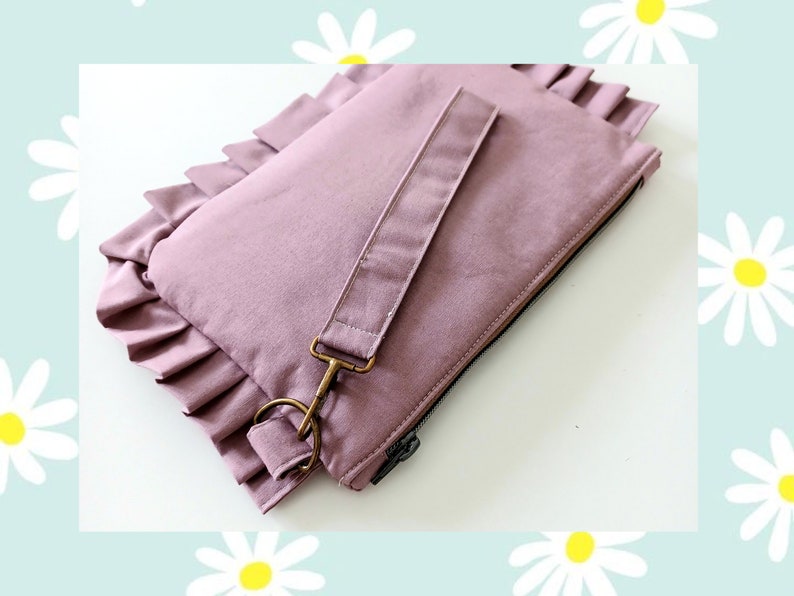 May include: A mauve fabric clutch bag featuring a ruffled edge detail along the side. The rectangular pouch includes a matching detachable wrist strap secured with a bronze-toned metal lobster clasp and a black zipper closure along the top edge.