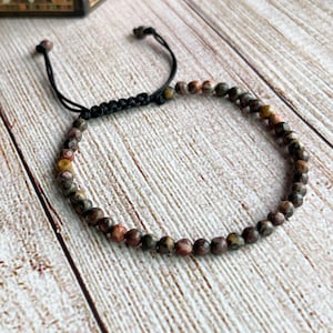 May include: A beaded bracelet featuring small, round, speckled beads in shades of brown, tan, and red. The bracelet has a black, adjustable macrame closure. The bracelet is displayed on a light wood surface.