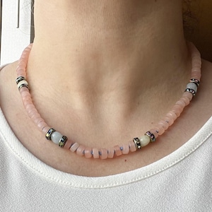 May include: A pink beaded necklace with alternating sections of small, square-cut beads and decorative spacers. The spacers are silver-toned with colorful accents and a central white bead. The necklace is worn against a white shirt.