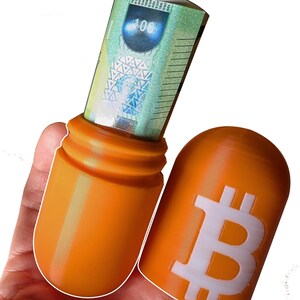 Puede incluir: Una cápsula de plástico naranja con un símbolo de Bitcoin blanco en un lado y un billete de 100 euros enrollado en el otro.