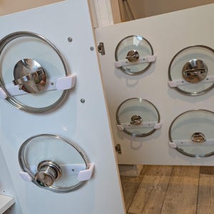 Könnte beinhalten: Weiße Schranktür mit mehreren Deckeln aus klarem Glas mit Edelstahlrändern und Griffen. Die Deckel werden von weißen Kunststoffhaltern gehalten. Die Schranktür ist geöffnet und zeigt weitere Deckel.