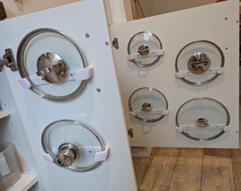 Op de afbeelding: Witte kastdeur met verschillende glazen pannendeksels met roestvrijstalen randen en handvatten. De deksels worden op hun plaats gehouden door witte plastic houders. De kastdeur is open en onthult meer deksels.