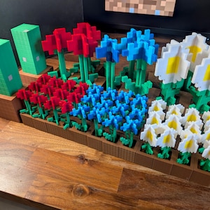May include: A wooden planter box filled with pixelated flowers in red, blue, and white, with green stems and leaves. Two green block structures are on the left. The flowers are arranged in rows, creating a colourful and unique garden display.