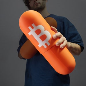 Può includere: Una persona che tiene un grande oggetto di forma di pillola arancione con un simbolo Bitcoin bianco su di esso.