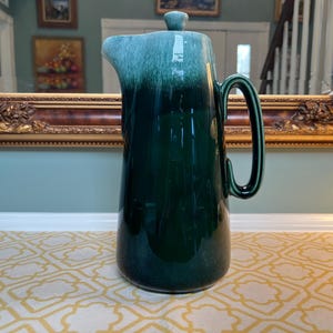 May include: A tall ceramic pitcher with a glossy finish, featuring a deep forest green base that transitions into a lighter, mottled teal glaze near the top rim. The pitcher includes a curved handle, a wide pouring spout, and a matching lid.