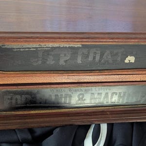 May include: A two-drawer wooden storage box with brass handles. The drawers have black labels with faded white text. The top label reads "J & P COAT". The bottom label reads "FOR HAND & MACH".