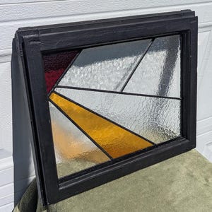 May include: A stained glass window with a black wooden frame. The glass features geometric shapes in red, yellow, white, and clear textured glass. The window is designed with angled lines and shapes, creating an abstract pattern.