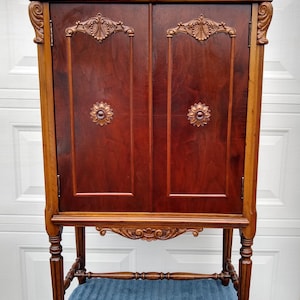 May include: A tall, antique wooden cabinet with two doors. The cabinet is a rich, dark reddish-brown color with lighter wood accents. It features ornate carvings on the doors, top, and legs, and is supported by four sturdy legs.