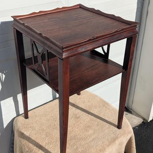 May include: A dark brown wooden side table with a square top and a lower shelf. The table has four slender legs and decorative cutouts on the sides. The table is approximately 24 inches tall.