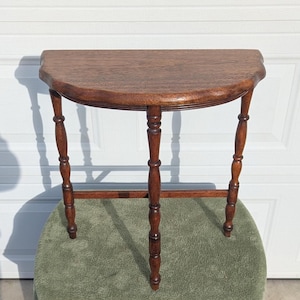 May include: A half-circle wooden table with a dark brown finish. The table has three turned legs and a lower stretcher for stability. The table is approximately 30 inches wide and 15 inches deep.