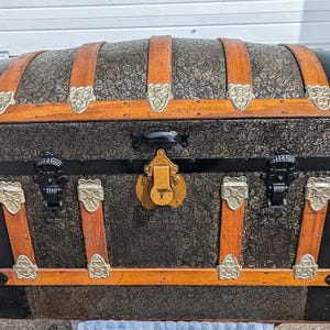May include: A vintage, black and brown, wooden travel trunk with metal accents and a brass lock. The trunk has a patterned design on the front and sides.