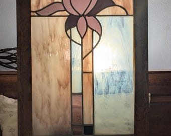 May include: A rectangular stained glass panel framed in dark wood, featuring a central stylized pink flower design. The glass consists of translucent white, tan, and light blue panels with dark lead lines separating the geometric and floral shapes.