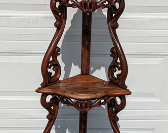 May include: A three-tiered corner shelf made of dark wood with intricate carvings. The shelf has a triangular shape and features a decorative finial on top.