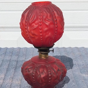 May include: A vintage red glass oil lamp with a round, textured shade and base. The lamp features a brass burner and a decorative, embossed pattern. The shade and base are a deep red color, and the lamp is set against a neutral background.