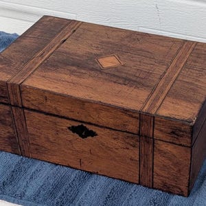 May include: A rectangular wooden box with a dark brown finish and visible wood grain. The lid features a centered diamond-shaped inlay and two vertical decorative bands. A black metal keyhole escutcheon is centered on the front panel.
