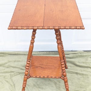 May include: A vintage wooden side table with a square top and a lower shelf. The table is made of light brown wood and features decorative carved edges and turned legs. The table measures approximately 24 inches tall.