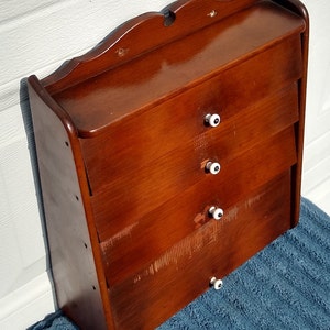 Puede incluir: Un armario de madera de pared con cuatro cajones y pomos de porcelana blanca. El armario es de color marr&oacute;n oscuro y tiene una parte superior ligeramente curvada.
