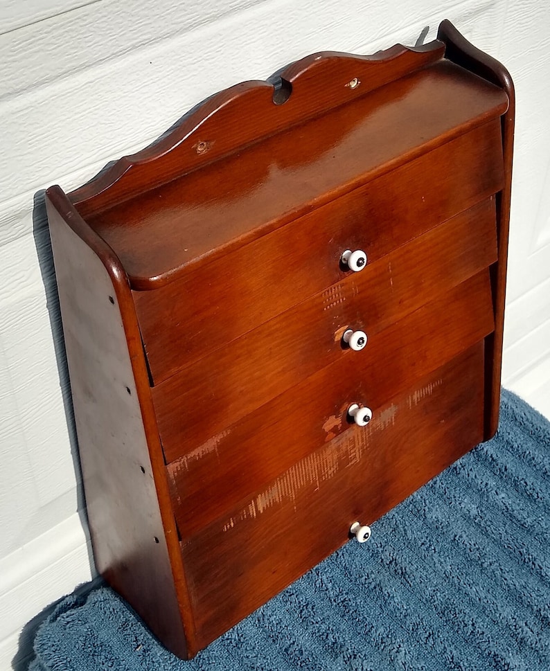 Puede incluir: Un armario de madera de pared con cuatro cajones y pomos de porcelana blanca. El armario es de color marr&oacute;n oscuro y tiene una parte superior ligeramente curvada.