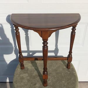 May include: A half-moon shaped wooden table with a dark brown finish. The table has three turned legs and a lower stretcher for stability. The table is approximately 30 inches wide.