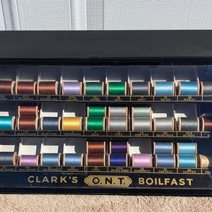 May include: A vintage wooden display case with rows of spools of thread in various colors. The case has a black background and a glass front. The label on the case reads "Clark's O.N.T. Boilfast".