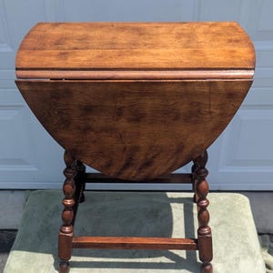 May include: A brown wooden drop-leaf table featuring a rectangular top with rounded edges and two hinged side leaves. The table is supported by four turned legs with decorative bulbous detailing and connected by horizontal stretchers near the base.