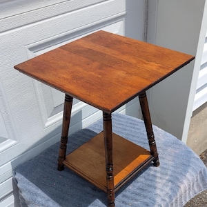 May include: A small, square, wooden side table with a warm brown finish. The table has a top surface of approximately 24 inches by 24 inches, supported by four turned legs and a lower shelf. The table is likely vintage or antique.