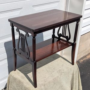 May include: A dark brown wooden side table with a rectangular top and a lower shelf. The table features four legs with vertical fluting and two decorative lyre-shaped supports on the sides, connecting the upper and lower sections of the frame.
