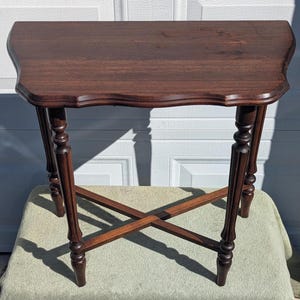 May include: A dark brown wooden side table featuring a rectangular top with curved, scalloped edges. The table is supported by four slender, turned legs that are connected by a central X-shaped cross-stretcher base for structural stability.