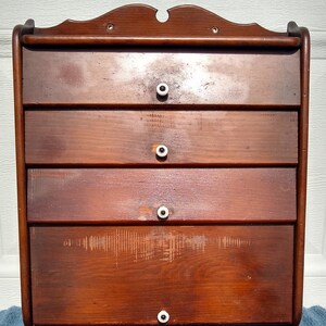 Puede incluir: Un armario de pared de madera con cuatro cajones y pomos de porcelana blanca. El armario tiene un tinte marr&oacute;n oscuro y un acabado ligeramente desgastado.