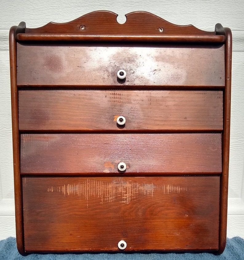 Puede incluir: Un armario de pared de madera con cuatro cajones y pomos de porcelana blanca. El armario tiene un tinte marr&oacute;n oscuro y un acabado ligeramente desgastado.