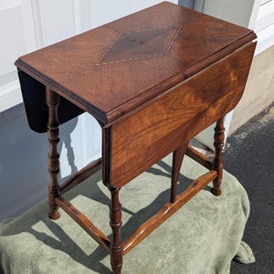 May include: A brown wooden drop-leaf table featuring a rectangular top with a central diamond-shaped inlay pattern. The table is supported by four turned legs connected by curved stretchers, showcasing a polished wood grain finish throughout.