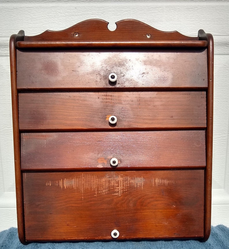 Puede incluir: Un armario de pared de madera con cuatro cajones y pomos de porcelana blanca. El armario tiene una parte superior ligeramente curvada y est&aacute; hecho de madera oscura.