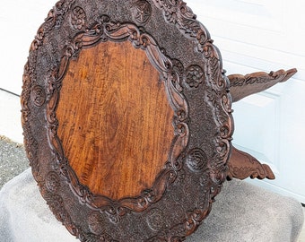May include: A round, intricately carved wooden table with a dark brown finish. The table top features a raised, ornate border with floral and geometric designs. The table legs are also carved with intricate details.
