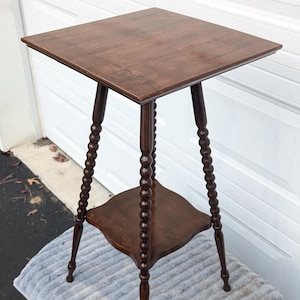 May include: A dark brown, square-top side table with a lower shelf. The table has three legs with a turned, beaded design. The table is approximately 28 inches tall.