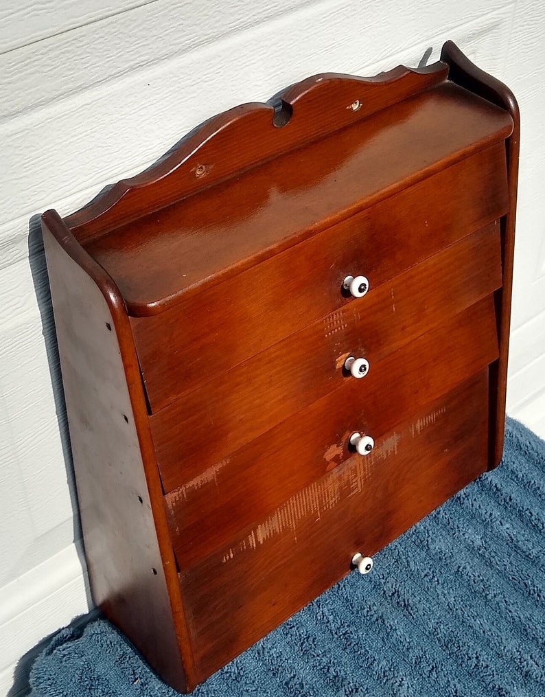 Puede incluir: Un armario de madera de pared con cuatro cajones y pomos de porcelana blanca. El armario tiene un tinte marr&oacute;n oscuro y un borde superior decorativo.
