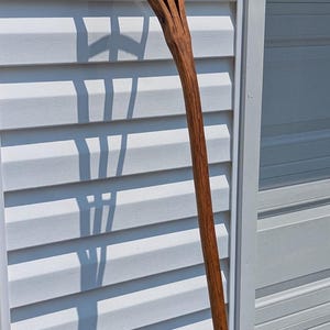 May include: A handcrafted wooden pitchfork with four tines. The long, curved handle and tines are a rich brown color. The pitchfork is leaning against a white wall, casting a shadow on a blue towel.