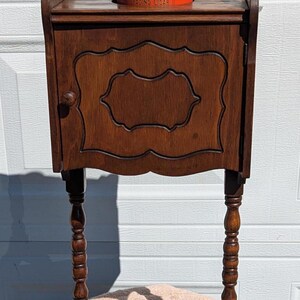 May include: A dark wood side table with a single door and a shelf. The table has turned legs and a small shelf on top. A red tin with the words "Traveler Leading Cigar" is sitting on the top shelf.
