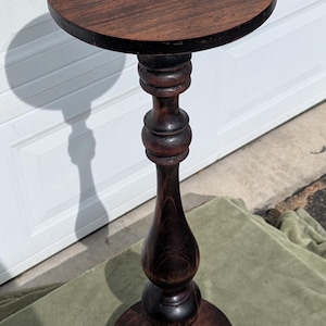 May include: A dark brown, antique-style plant stand with a round top and base. The stand is approximately 30 inches tall and features a turned, baluster-shaped central column. The wood grain is visible throughout the stand.