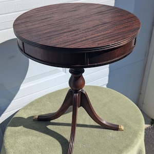 May include: A dark brown, round side table with a pedestal base and three legs. The tabletop has a patterned wood grain. The legs have gold-colored accents. The table is approximately 24 inches in diameter.