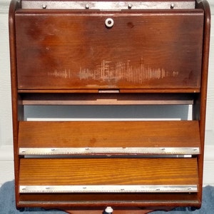 Puede incluir: Una estanter&iacute;a de pared de madera con dos estantes y una puerta abatible. La estanter&iacute;a tiene un acabado marr&oacute;n claro y un ribete de metal plateado. La puerta tiene un pomo blanco.