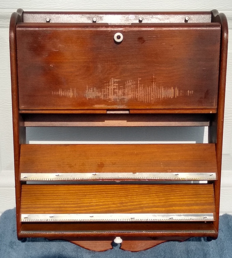 Puede incluir: Una estanter&iacute;a de pared de madera con dos estantes y una puerta abatible. La estanter&iacute;a tiene un acabado marr&oacute;n claro y un ribete de metal plateado. La puerta tiene un pomo blanco.