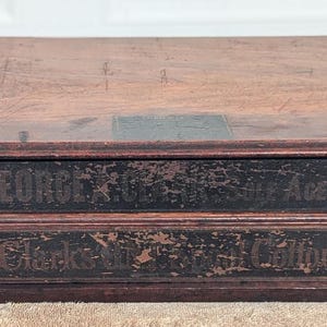 May include: A vintage wooden box with two drawers and a hinged lid. The box is stained a dark brown color, with white knobs on the drawers. The text "GEORGE S. AGENT" and "Jack's M. & Cotton" are visible on the drawers.