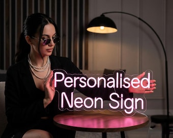 May include: A neon sign with the words "Personalised Neon Sign" in a pink glow. The sign is held by a person. The background includes a lamp and a wooden table.