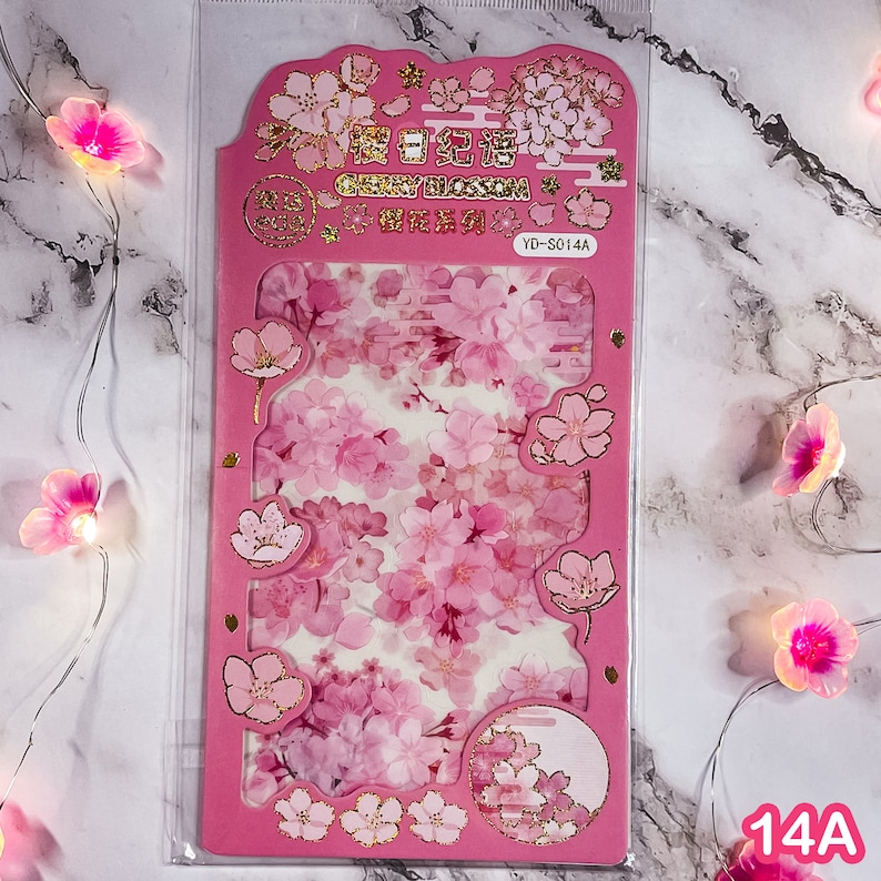 Pu&ograve; includere: Un foglio di adesivi rosa Cherry Blossom con dettagli dorati. Il foglio presenta vari disegni di fiori di ciliegio e il testo "Cherry Blossom" in inglese e caratteri cinesi. Il foglio di adesivi &egrave; etichettato "YD-S014A".