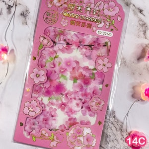 Pu&ograve; includere: Una confezione rosa di adesivi di fiori di ciliegio. Gli adesivi presentano varie tonalit&agrave; di fiori rosa con dettagli dorati. La confezione ha forme ritagliate e testo in oro e nero. Gli adesivi sono in una custodia di plastica trasparente.